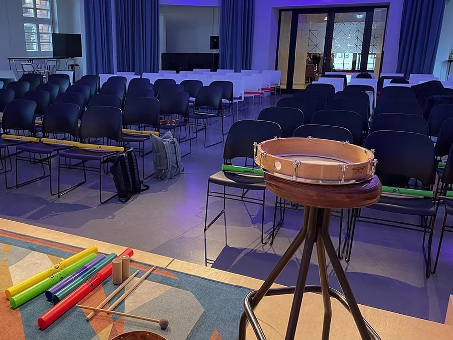 Trommel auf einem Ständer mit verschiedenen Musikinstrumenten im Vordergrund und leeren Stühlen im Hintergrund.
