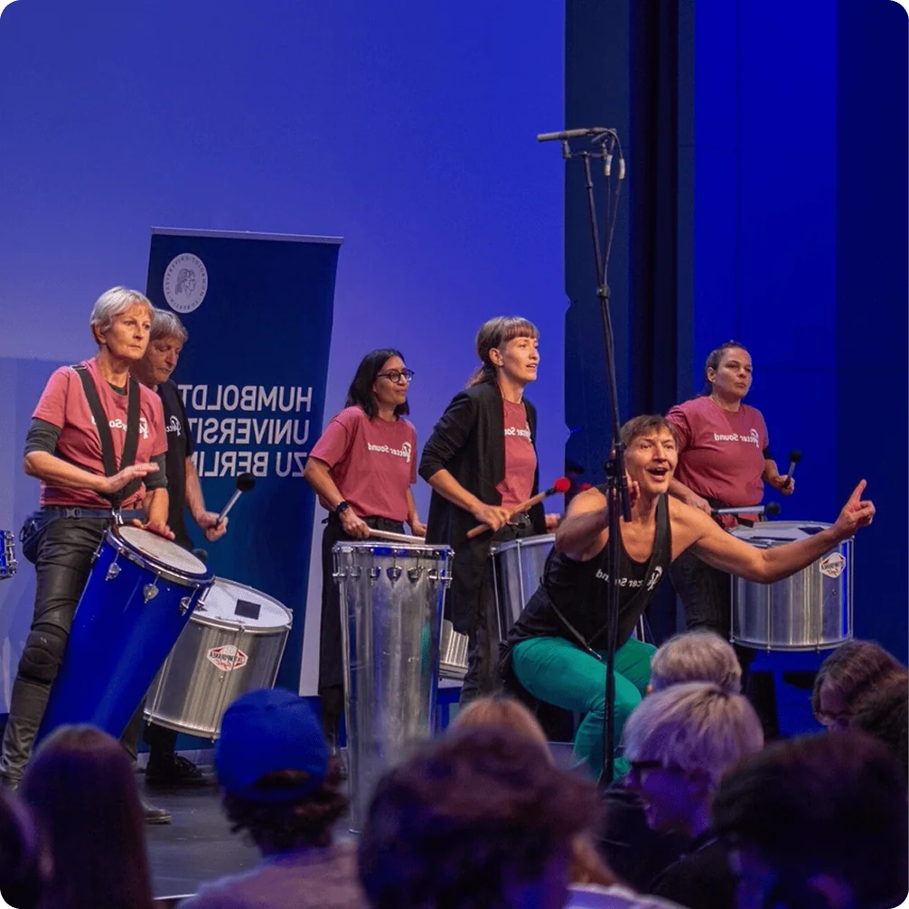 Gruppe von Musikerinnen mit Percussion-Instrumenten auf einer Bühne während einer Aufführung. Sie tragen einheitliche T-Shirts und spielen verschiedene Trommeln.